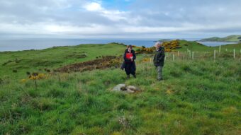 Two people standing in a field near a rock in the ground by water